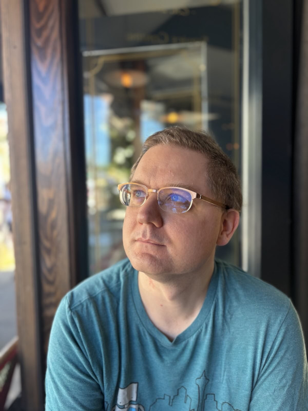 John Ayliff, a white guy with short blond hair, blue eyes, bronze-rimmed glasses, and a blue T-shirt, pictured here in a cafe on a sunny day gazing up and to one side as if daydreaming about space.